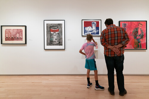 An adult and a child stand with their backs to us, viewing five artworks hanging on a wall